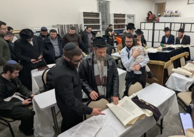 Men gathered around a table as an open scroll is read, with others looking on and one holding a sleeping child