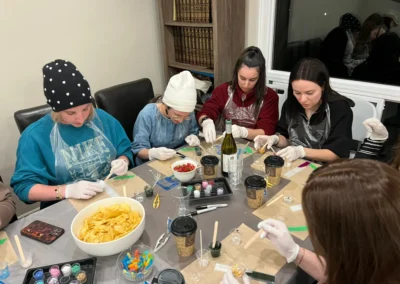 Four women in aprons and gloves working on a craft project at a table with coffee cups and snacks