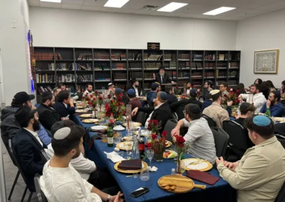 A speaker at a lectern addresses guests seated at long tables with navy tablecloths and red roses, in front of bookshelves
