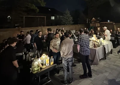 An evening gathering on a backyard patio, with people standing around tables lit by small lamps