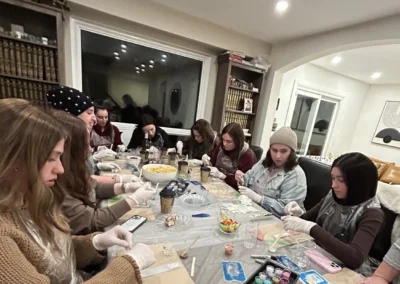 Women in aprons and gloves seated along a long table doing a craft activity