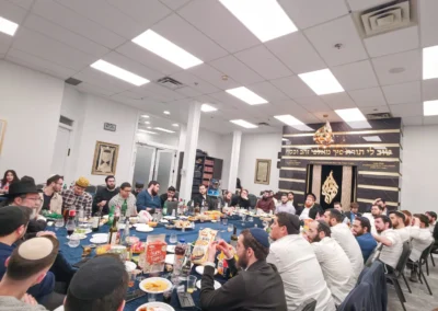 A large group seated around a long table in a hall with a black-and-gold ark and Hebrew lettering