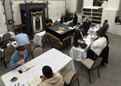 Men studying from open books at tables in a shul room with an ark and a velvet-covered bimah