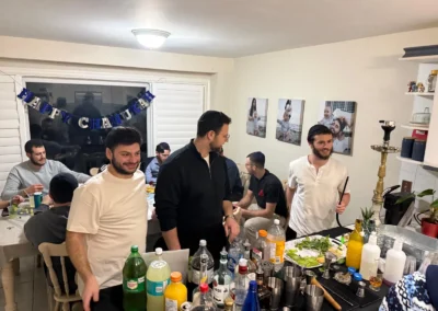 Young men around a drinks table in a home, with a Happy Chanukah banner in the window