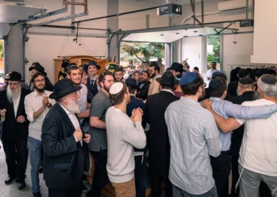 A crowded room of men dancing and clapping, with Torah scrolls in decorated covers in the background