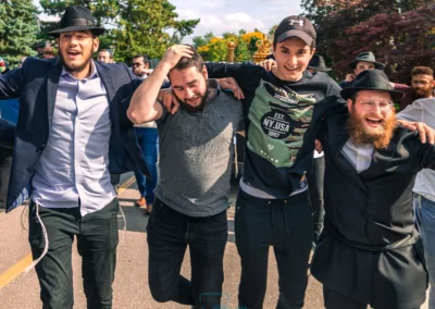 Four men dancing arm in arm outdoors on a sunny street lined with trees