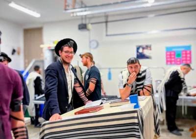 Men wearing tefillin and tallitot around a table draped with a striped tallit, one smiling at the camera