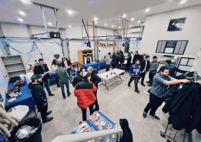 People mingling in a room decorated with blue streamers and a Happy Chanukah banner, with doughnuts on a table in the foreground